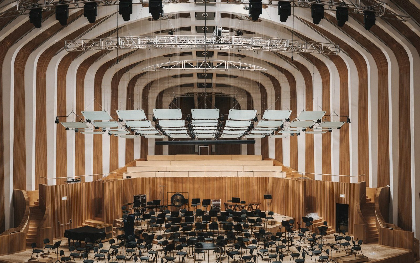 Empty concert hall stage with curved wooden ribbed ceiling and suspended acoustic panels above rows of chairs