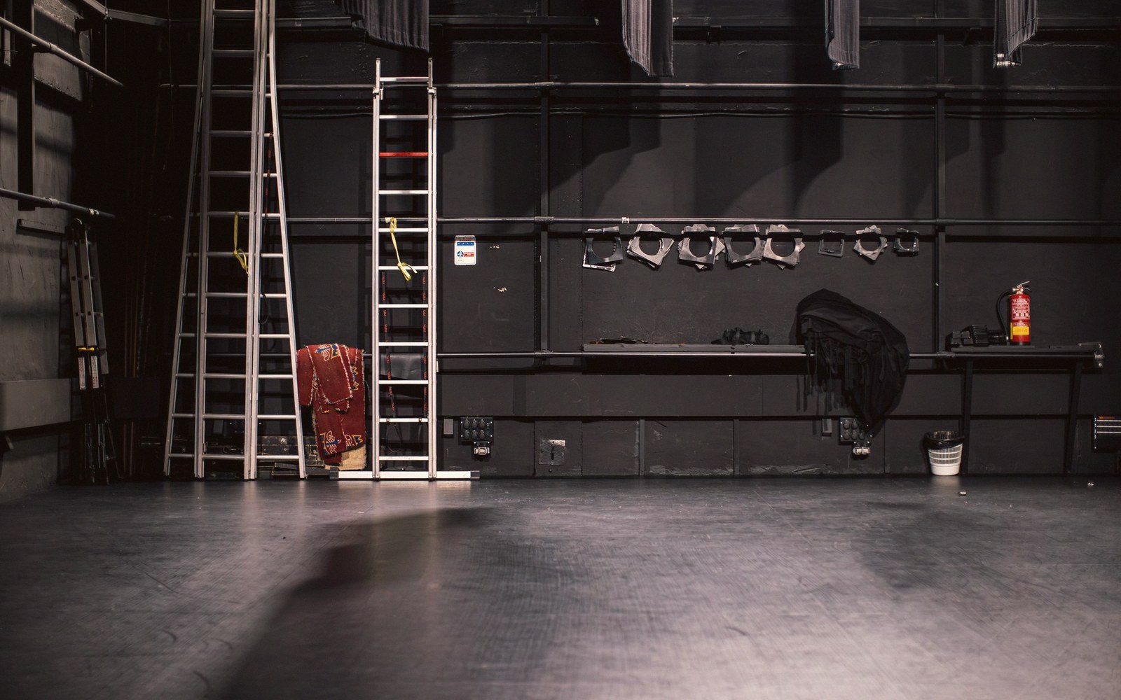 A painted stage set partially struck, bare scaffolding visible at one edge, a single work light casting hard shadow across the flats